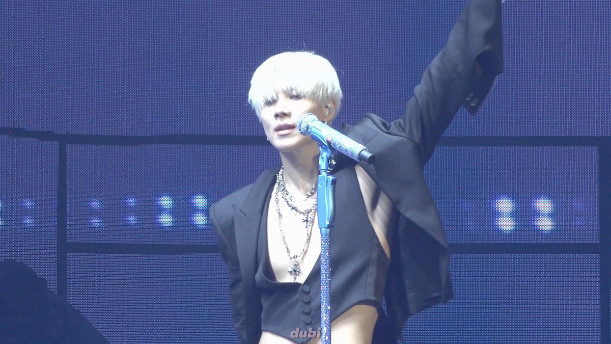 A performer with platinum blonde hair, wearing a black open suit jacket and layered silver cross necklaces, sings into a sparkly microphone on stage with one arm raised, with the watermark 'dubi' visible at the bottom.