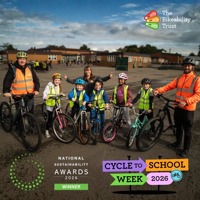 Group of children and adults wearing helmets and high-visibility vests posing with bicycles on a school playground under a cloudy sky.