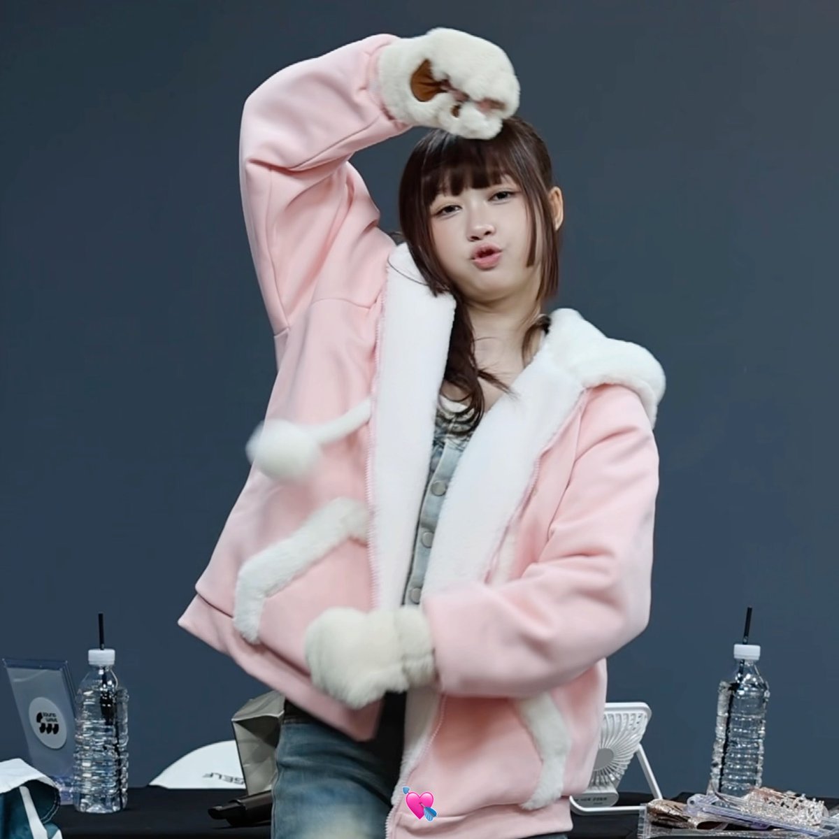 A young woman with brown hair and bangs wears a pink fleece jacket with white trim, posing with her arm raised above her head and making a kissy face.