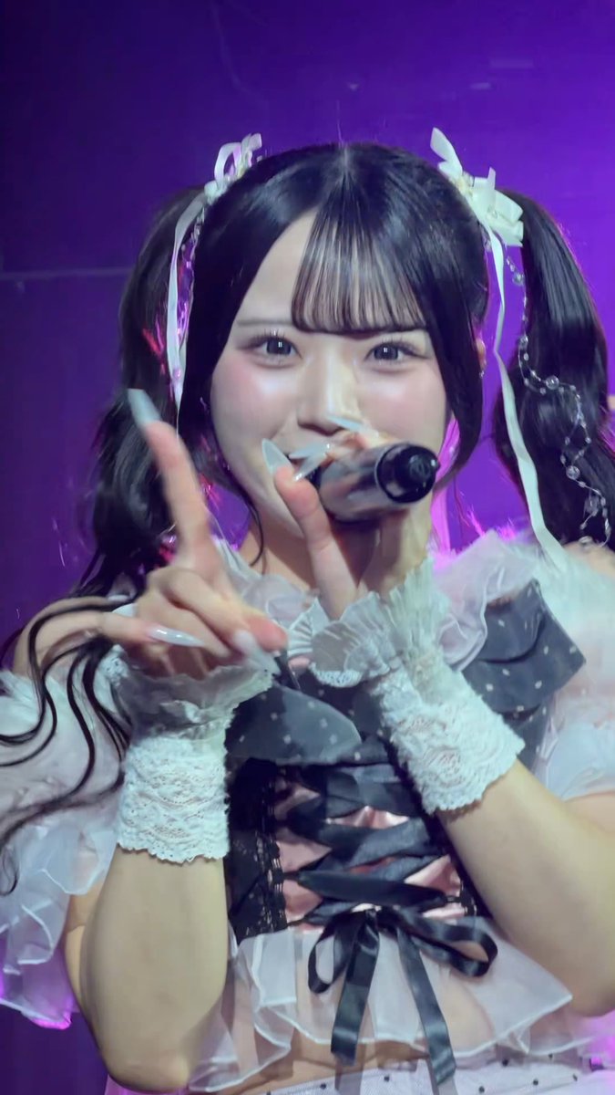 A young performer in a frilly white and black outfit with twin-tail hair ribbons sings into a microphone on stage under purple lighting, making a peace sign with her hand.