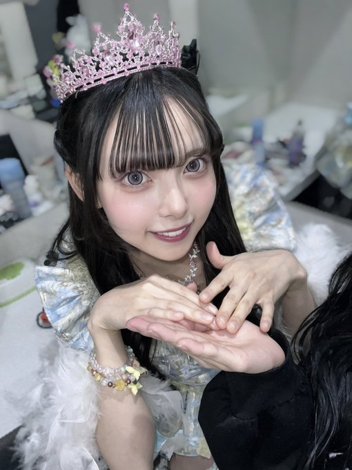 A selfie of a young woman with long dark hair and bangs, wearing a sparkling pink tiara, a decorative dress, and colorful charm bracelets, smiling at the camera with her hands forming a heart shape in front of her.
