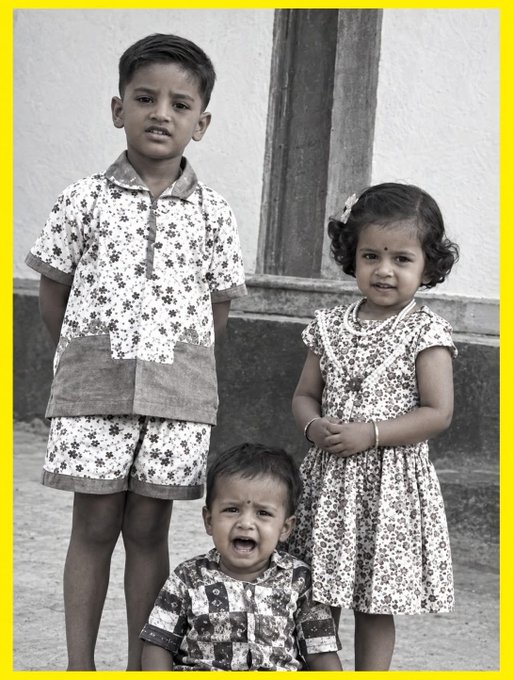 A black-and-white photograph showing three children—a boy in a patterned outfit standing on the left, a younger child sitting and crying in the middle, and a girl in a floral dress standing on the right—posed outdoors.