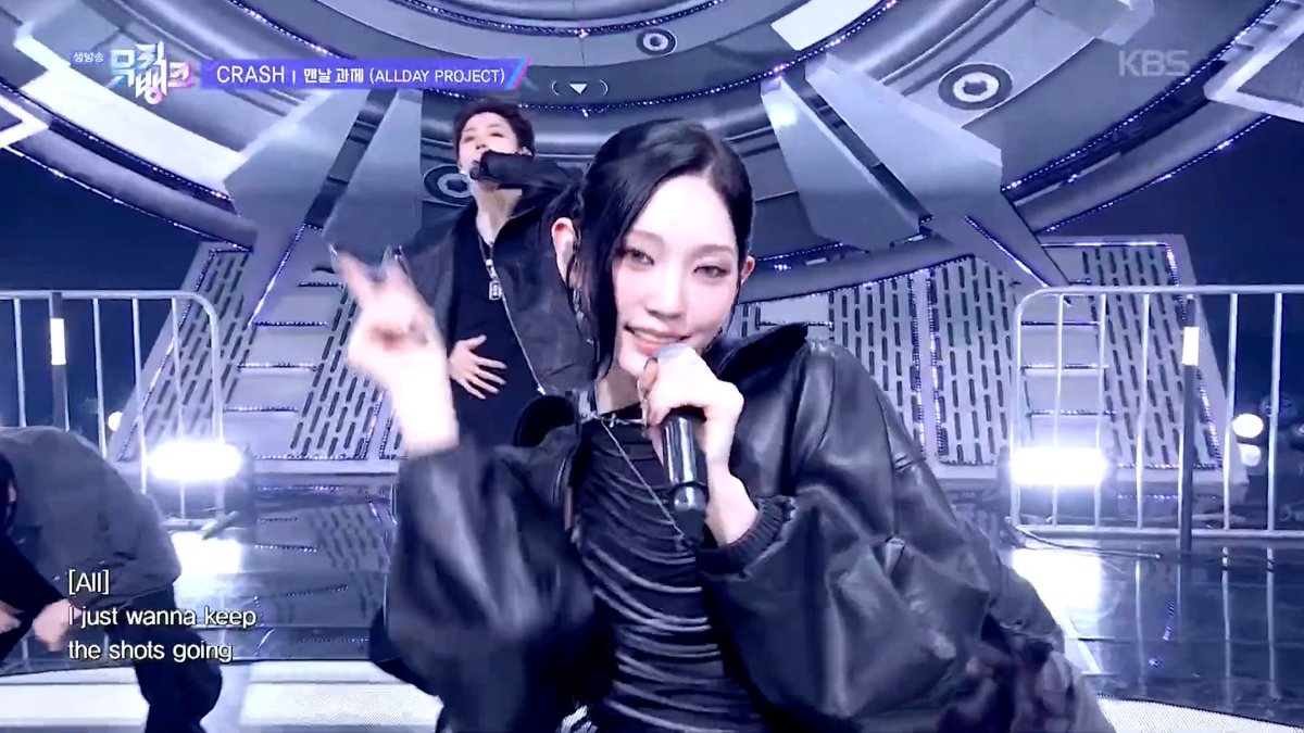 A KBS Music Bank performance of 'CRASH' by ALLDAY PROJECT, featuring a female singer with a microphone in the foreground and lyrics reading '[All] I just wanna keep the shots going'.