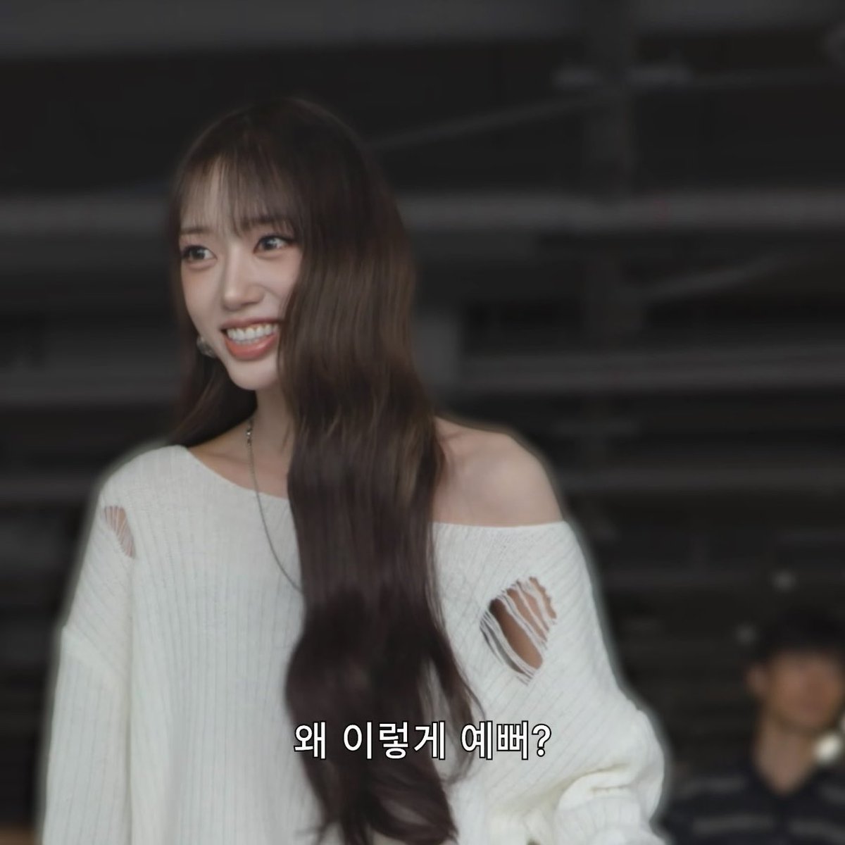 A smiling young woman with long brown hair and bangs, wearing a distressed white off-shoulder knit sweater, is shown against a dark industrial background. Korean text at the bottom reads "왜 이렇게 예뻐?" (meaning "Why are you so pretty?"), matching the post's caption as she gazes off to the side with a bright, cheerful smile that seems to embody the sentiment of her own compliment.