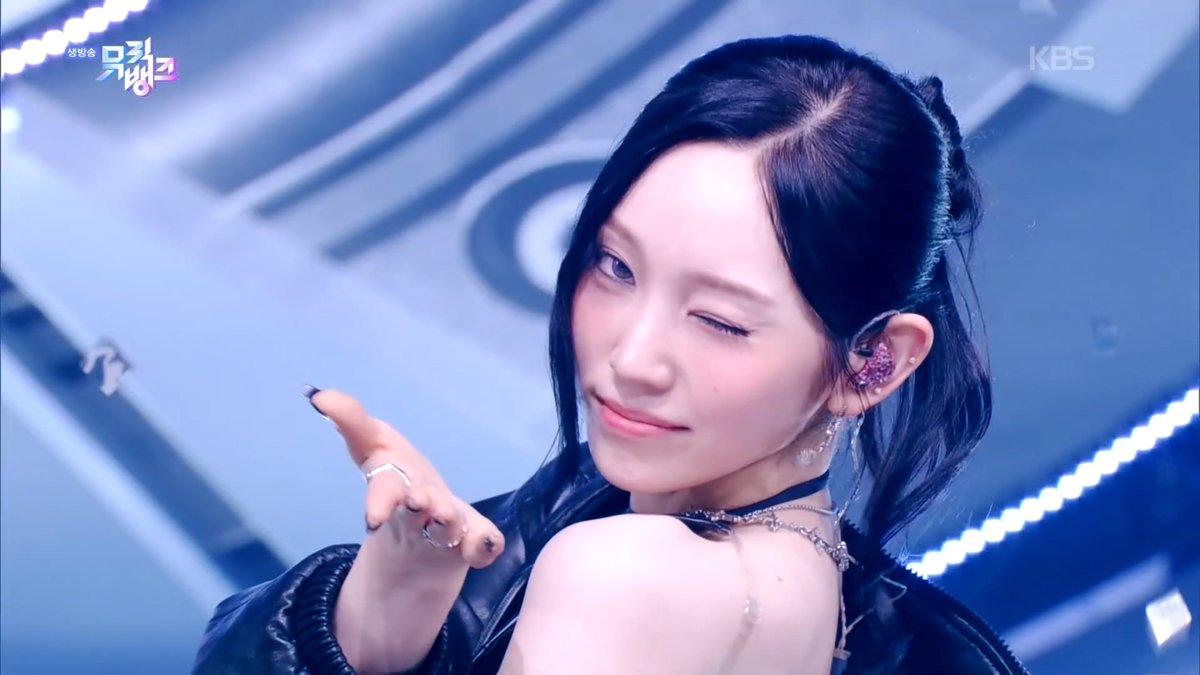 A K-pop performer on a brightly lit stage winks playfully at the camera while posing, with the KBS logo and the show title "뮤직뱅크" (Music Bank) visible in the corners.
