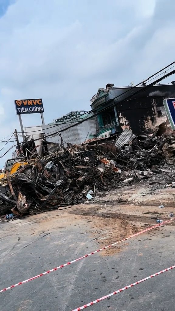 The aftermath of a fire at a commercial building, showing extensive rubble and debris piled against a damaged structure with a 'VNVC Tiêm Chủng' (vaccination clinic) sign visible, cordoned off with red and white safety tape.