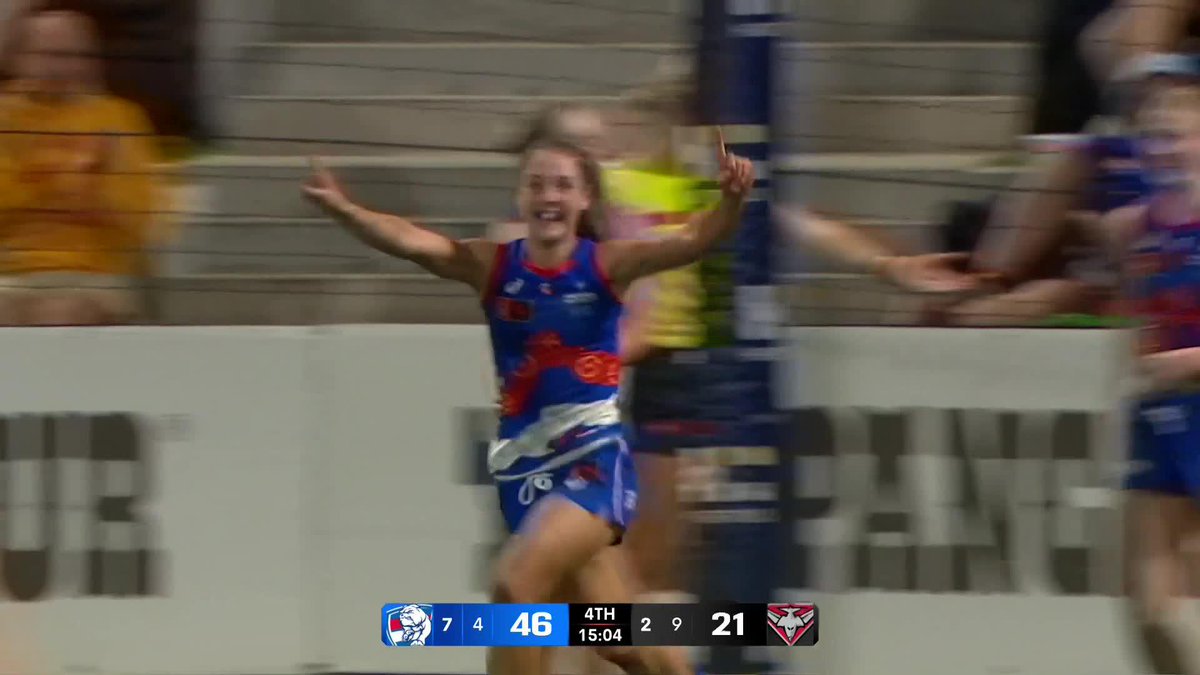 An action shot from an Australian Rules Football match showing a player in a blue and red uniform celebrating with arms raised, with a scoreboard at the bottom reading '7 4 46 4TH 15:04 2 9 21'.