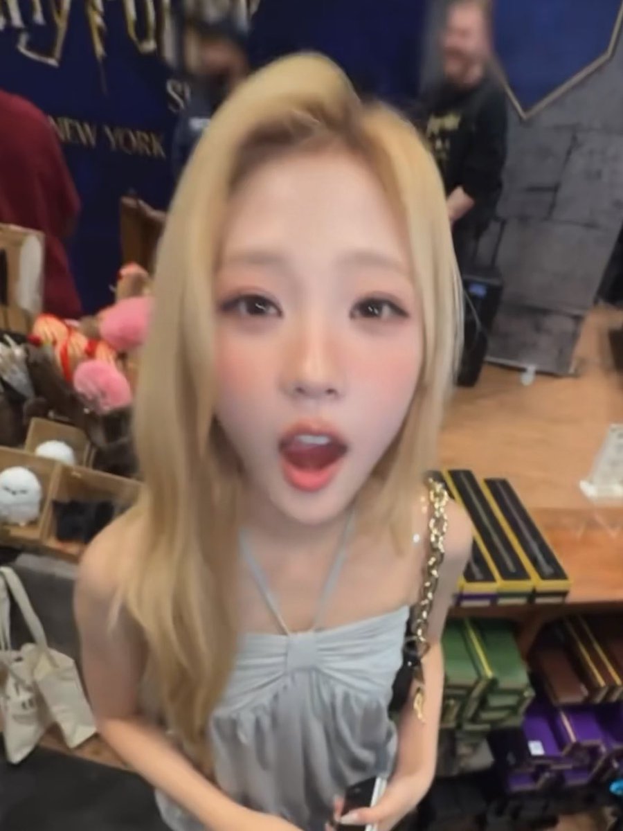 A close-up of a young woman with blonde hair and red lipstick in what appears to be a store, with boxes and stuffed toys visible in the background along with a 'NEW YORK' poster.