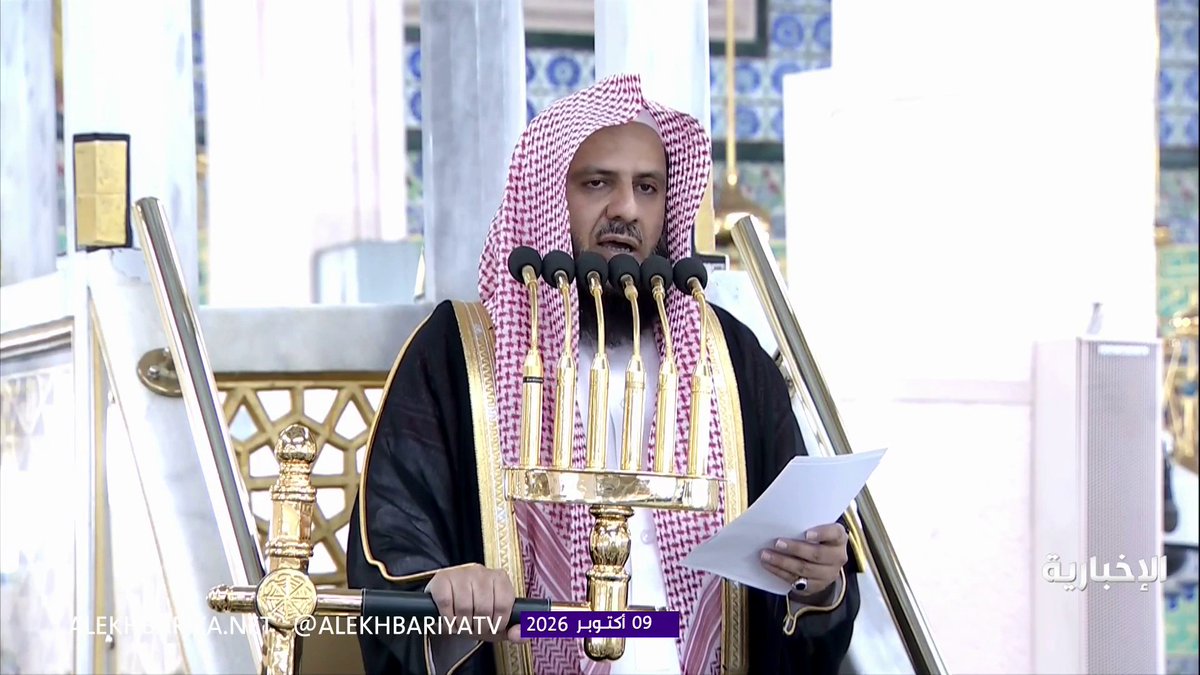 A man in traditional Arab attire (red and white keffiyeh with black bisht) delivers a sermon from an ornate golden pulpit, speaking into a cluster of five microphones while holding papers. The Al-Ekhbariya TV broadcast is dated October 9, 2026, and shows the Imam of Al-Masjid an-Nabawi warning of severe divine punishment for those who intend harm against Mecca, emphasizing Allah's protection of the holy city.