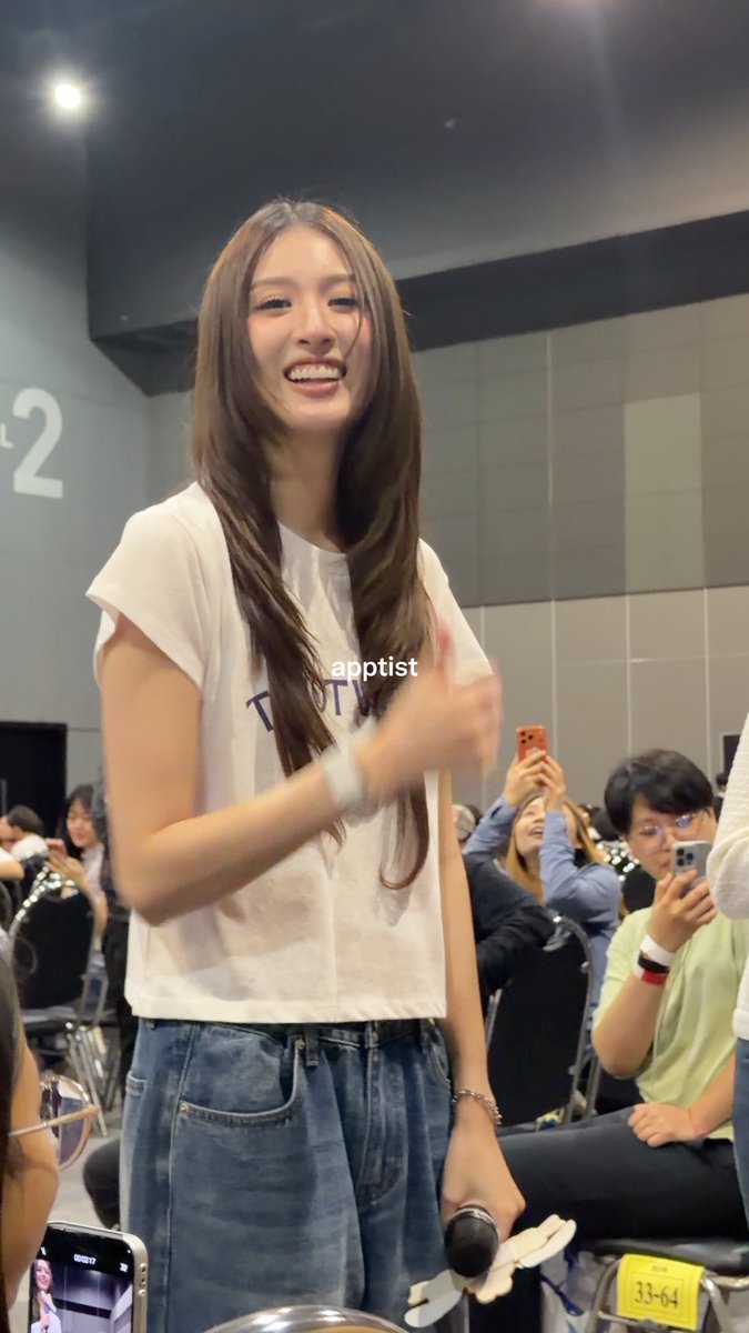 A young woman with long brown hair smiles while standing in front of an audience that is taking photos of her at what appears to be a fan event, with the watermark "apptist" visible on a white t-shirt and denim skirt she's wearing, matching the post's reference to the GMMTV Fanival 2026 event and her being a fan of View and Mim.