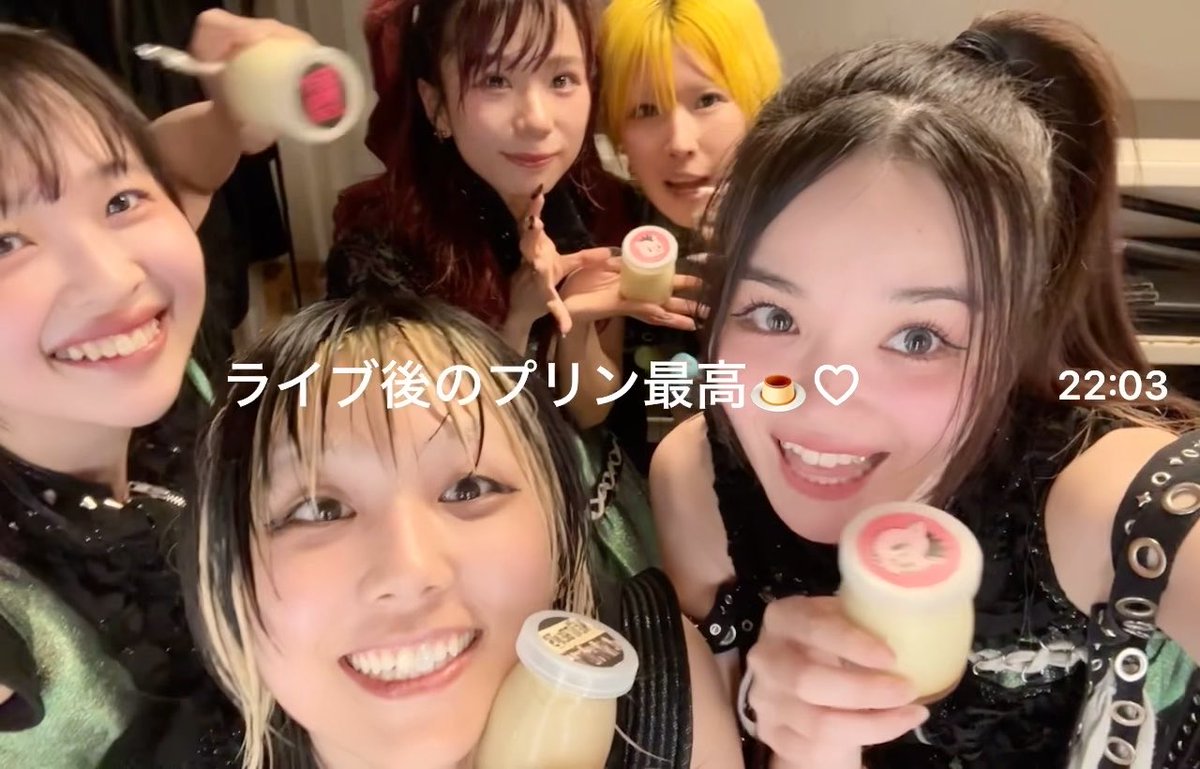 A group of five young women smile for a selfie while holding small pudding cups, with Japanese text overlay reading 'ライブ後のプリン最高🍮🤍' and a timestamp of '22:03'.