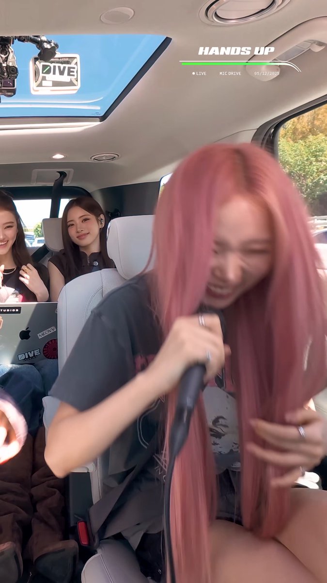 A pink-haired performer (likely Gawon) passionately sings into a microphone while laughing, with the "HANDS UP" broadcast title and "LIVE MIC DRIVE 05/12/2025" text visible at the top of the car-ride karaoke stream, while other group members smile from the backseat.