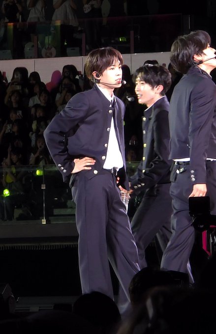 A young male performer stands center stage in a navy blue Japanese school uniform (gakuran) with matching trousers, wearing a headset microphone while performing alongside two other members dressed similarly. The image captures a live concert moment, with the audience visible in the background, matching the post's reference to the traditional Japanese school uniform style associated with the performance.