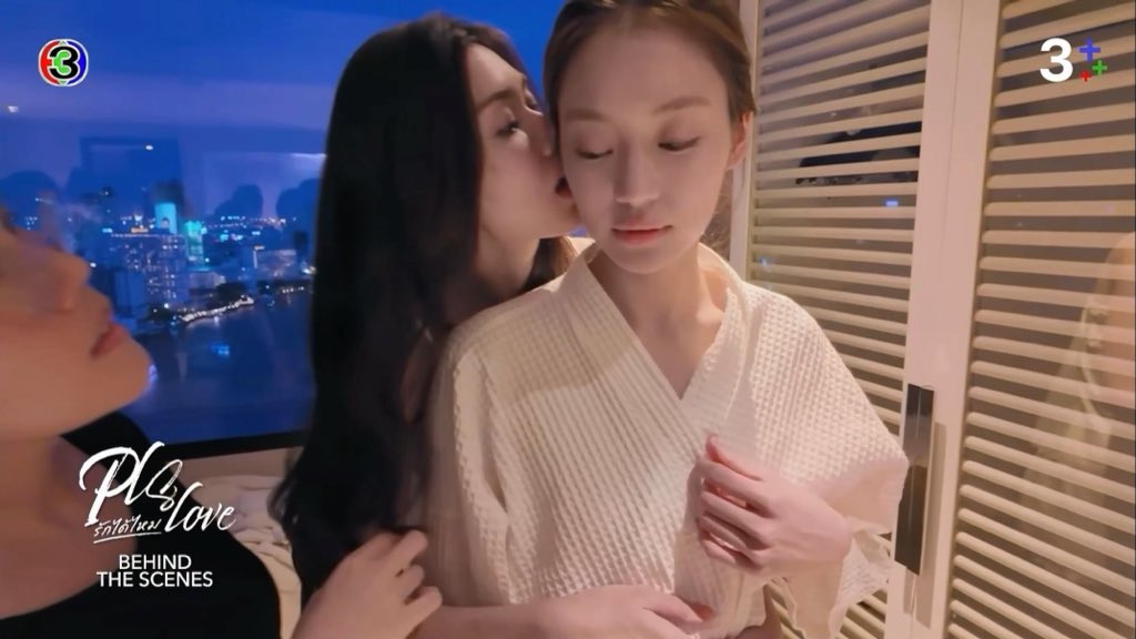 Two women share an intimate moment as one kisses the other's ear in a cozy setting with a city skyline visible in the background. The image is from "Behind the Scenes" content for "Pl.s. rak wiki ploen" (Secret of Love), as indicated by the logos and text in the corners, showing the playful behind-the-scenes interaction where the actresses joke about adding creative flair to their ear-nibbling scene.