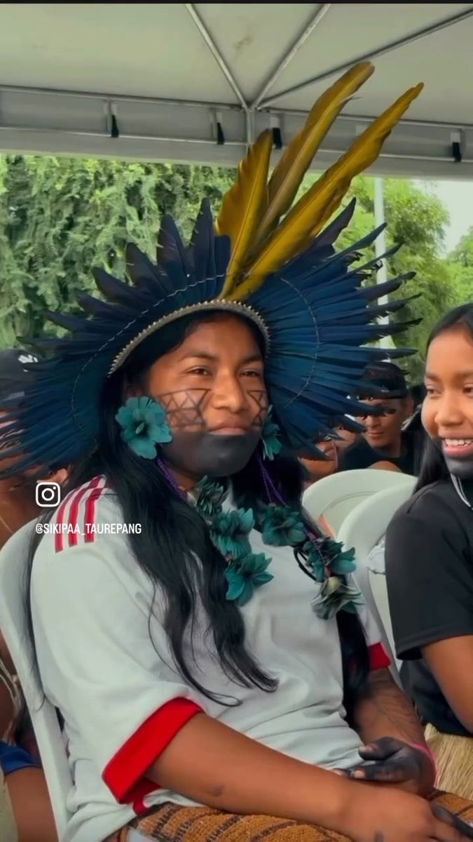 Photo of an Indigenous woman wearing traditional attire including a feathered headdress with yellow and blue feathers, face paint with geometric patterns, teal flower earrings, and a flower necklace, with the Instagram handle '@sikipaa_taurepang' visible.