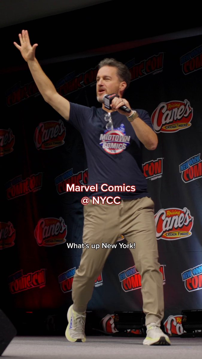 Man on stage at NYCC wearing a Midtown Comics shirt, holding a microphone and greeting the audience with a wave. The backdrop features NYCC and Raising Cane's logos, tying to the Marvel Comics panel teaser about the upcoming Midnight line.