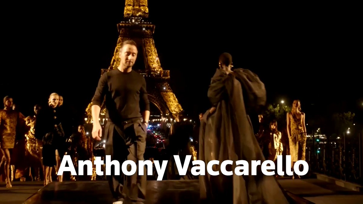 The image captures a nighttime fashion show scene at the base of the Eiffel Tower, featuring designer Anthony Vaccarello—identified by bold white on-screen text "Anthony Vaccarello" overlaid at the bottom—as he walks alongside models in dark, flowing garments, visually illustrating Kering's announcement of his departure from Saint Laurent after a decade at the helm.