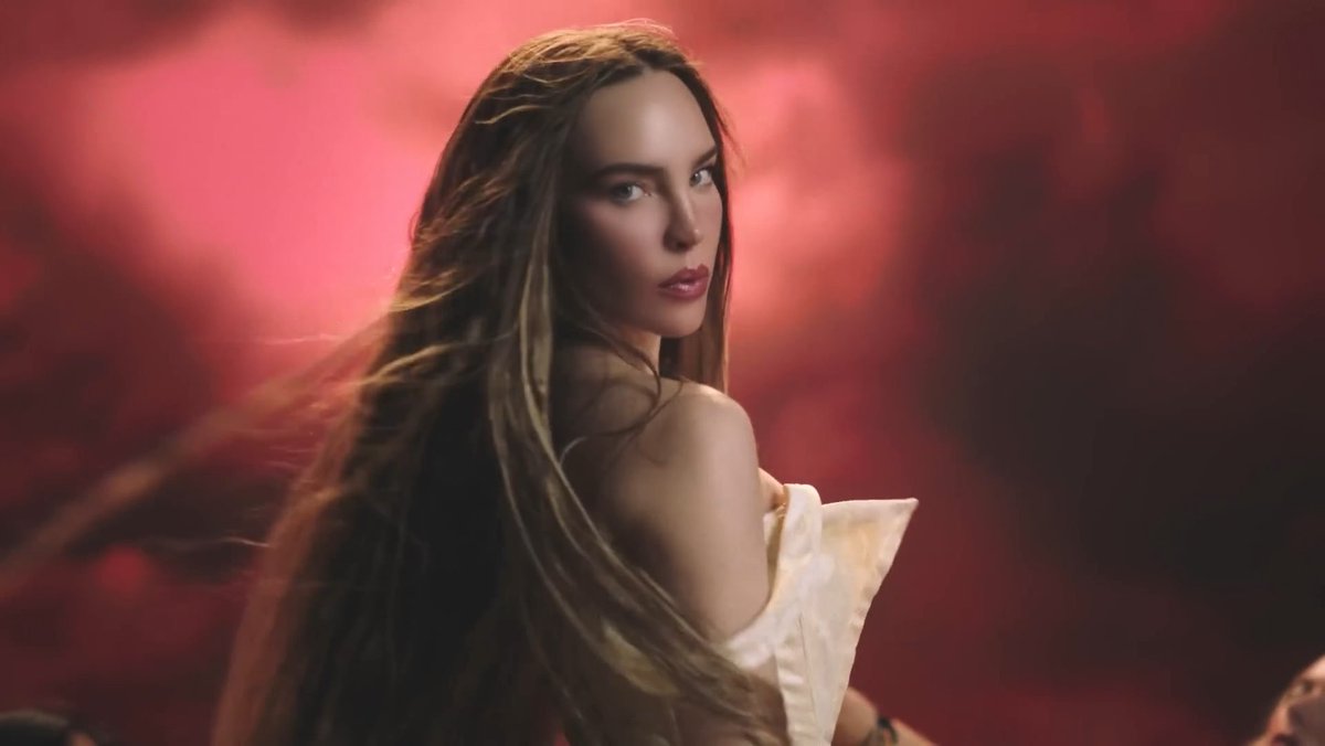 A woman with long, flowing brown hair looks over her shoulder against a dramatic, smoky red background, wearing a white off-the-shoulder garment.