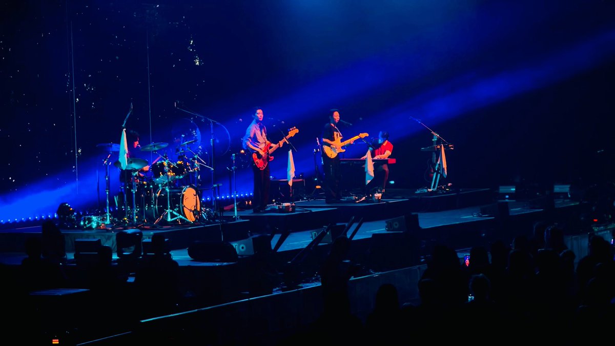 A band performs live on stage bathed in deep blue lighting, with visible members playing instruments including a bass, electric guitar, and keyboard, while an audience watches in the foreground.