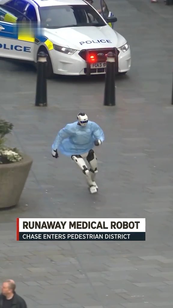 A humanoid medical robot in blue scrubs runs across a pedestrian plaza while being pursued by a British police car, with a news banner reading 'RUNAWAY MEDICAL ROBOT - CHASE ENTERS PEDESTRIAN DISTRICT'.