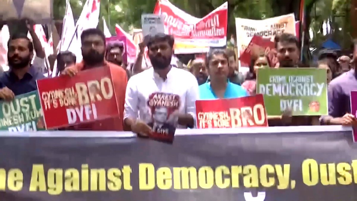 A protest rally with demonstrators holding signs reading 'OUST DYFI', 'CYUNESH', 'BRO DYFI', and 'ERINP AGAINST DEMOCRACY', with a banner below stating 'Against Democracy. Oust'.