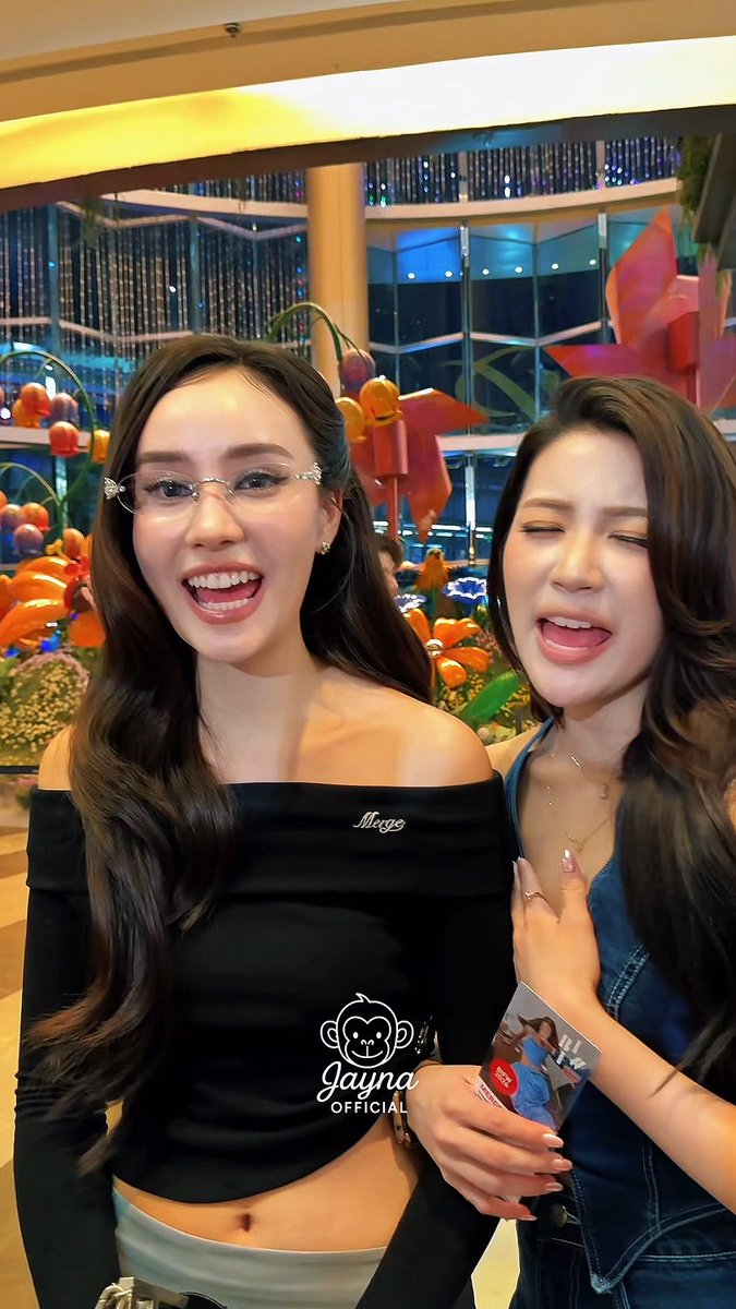 Two young women pose together; one wears glasses and a black off-the-shoulder top with 'Mege' embroidered on it, while the other wears a denim outfit and makes a playful face. The image has a 'Gayna Official' monkey logo watermark.