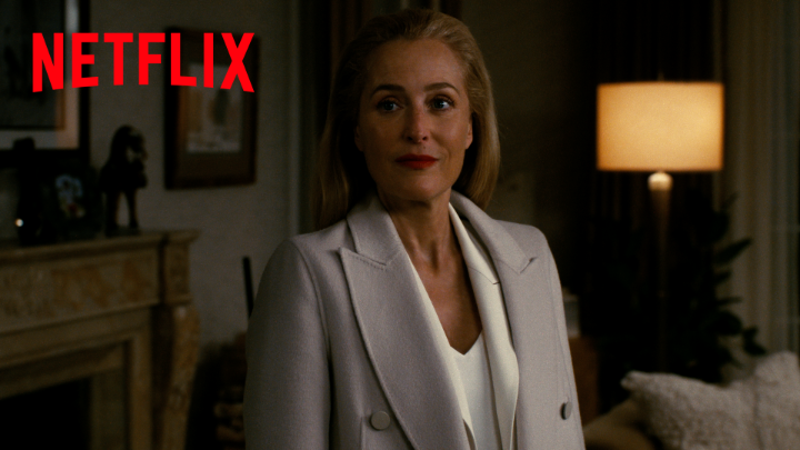 A woman with blonde hair wearing a light gray blazer over a white top stands in a dimly lit room, with the red 'NETFLIX' logo displayed in the upper left corner.
