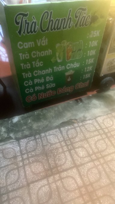 A photograph of a green signboard titled 'Trà Chanh Tắc' displaying a beverage menu in Vietnamese, including items like 'Cam Vắt' (25K), 'Trà Chanh' (10K), and 'Cà Phê Sữa' (15K), with a tiled floor visible below.