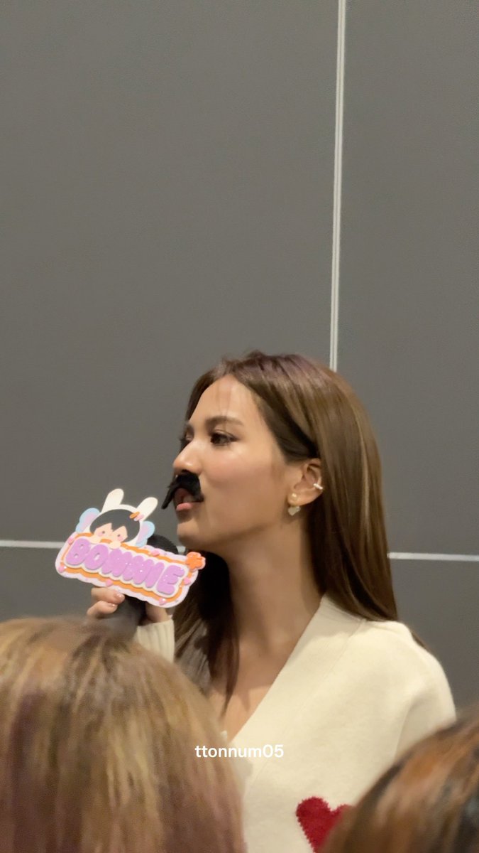 A woman wearing a white top with a red heart design speaks into a microphone while sporting a fake mustache, holding a sign that reads "BOMMIE."