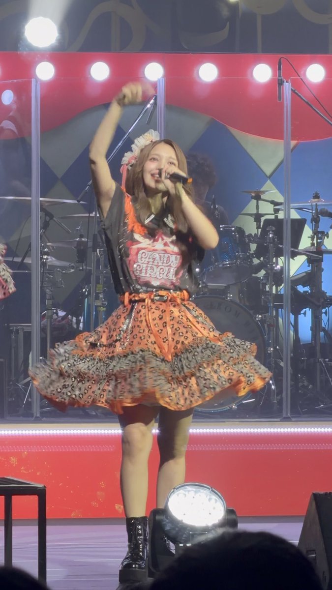 A performer on stage in an orange and black leopard-print dress holds a microphone and raises one arm, with a band playing behind her on a stage decorated with red banners.