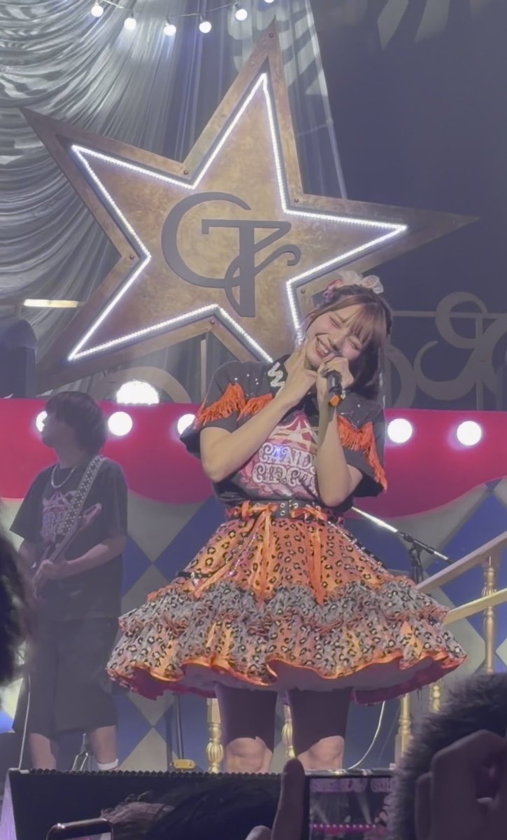 A Japanese idol performer in an elaborate orange, leopard-print, and black frilly outfit smiling and singing into a microphone on stage, with a guitarist visible and a large illuminated star featuring a 'CR' logo in the background.