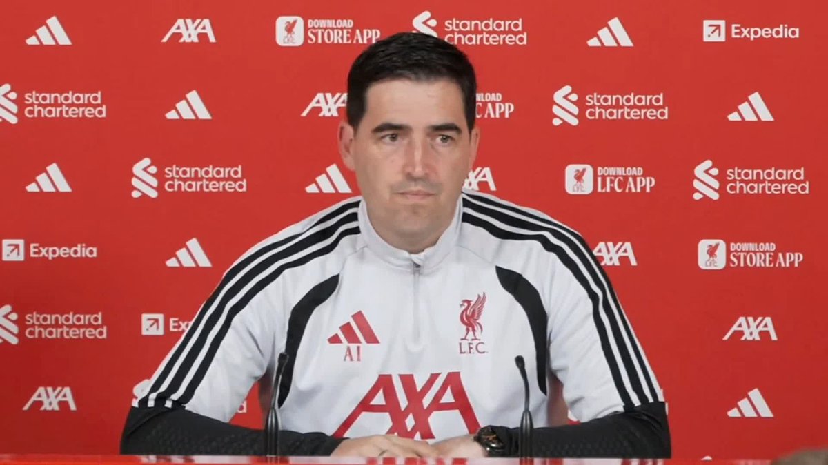 Andoni Iraola, wearing a white Liverpool FC training jacket with "AXA" and the LFC crest, sits at a press conference backdrop featuring sponsor logos including Standard Chartered, AXA, and Expedia. The image accompanies his quote expressing readiness to test his team's level against Manchester City.
