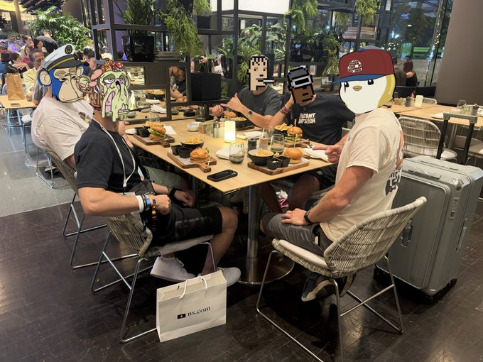 The image shows a group of friends (faces obscured by cartoon stickers) sharing a meal at a restaurant table filled with various dishes, drinks, and takeout bags, with one prominent bag displaying "ntucfairprice.com." A gray suitcase beside the table suggests recent travel, fitting the caption of enjoying dinner with friends after a long day in Singapore.