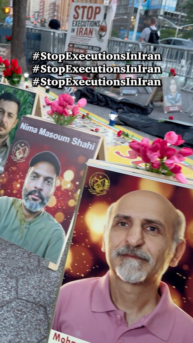 A memorial with framed portraits of individuals including one labeled 'Nima Masoum Shahi' and another beginning with 'Mohs...', set up with pink flowers and signs reading 'STOP EXECUTIONS IN IRAN' and overlaid with the hashtag '#StopExecutionsInIran'.