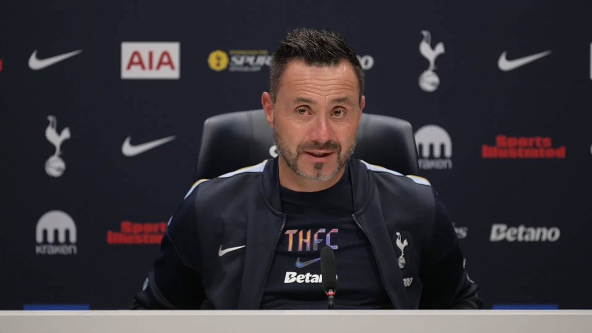 Roberto De Zerbi sits at a Tottenham Hotspur press conference, wearing a navy club jacket featuring the THFC crest and "Betano" sponsor logo, with backdrop displaying Tottenham's cockerel emblem, AIA, Kraken, Betano, and Sports Illustrated logos as he addresses the media regarding Dejan Kulusevski's fitness.