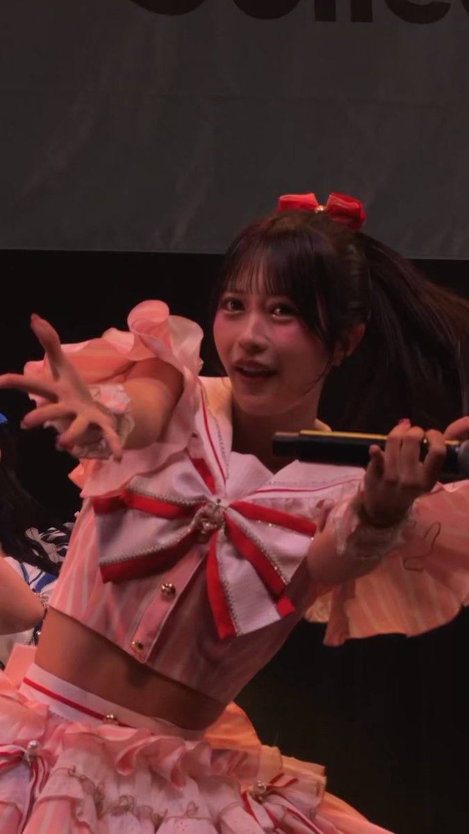 A young woman performs on stage in a pink and white idol costume with a large red and white bow, holding a microphone and reaching out toward the camera.