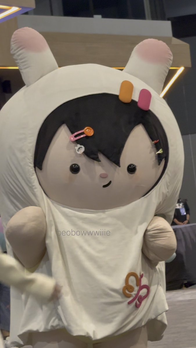 A mascot costume featuring a cartoon-style face with black hair adorned with colorful hair clips and pink-tipped bunny ears, with the username 'beobowwwiiie' visible on the white shirt.