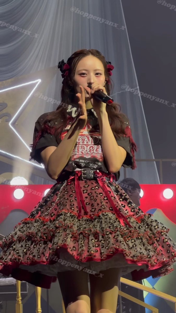 A concert photo of a performer in an elaborate red and black Gothic Lolita outfit with leopard print, singing into a microphone, watermarked with '@hoppenyanx_x'.