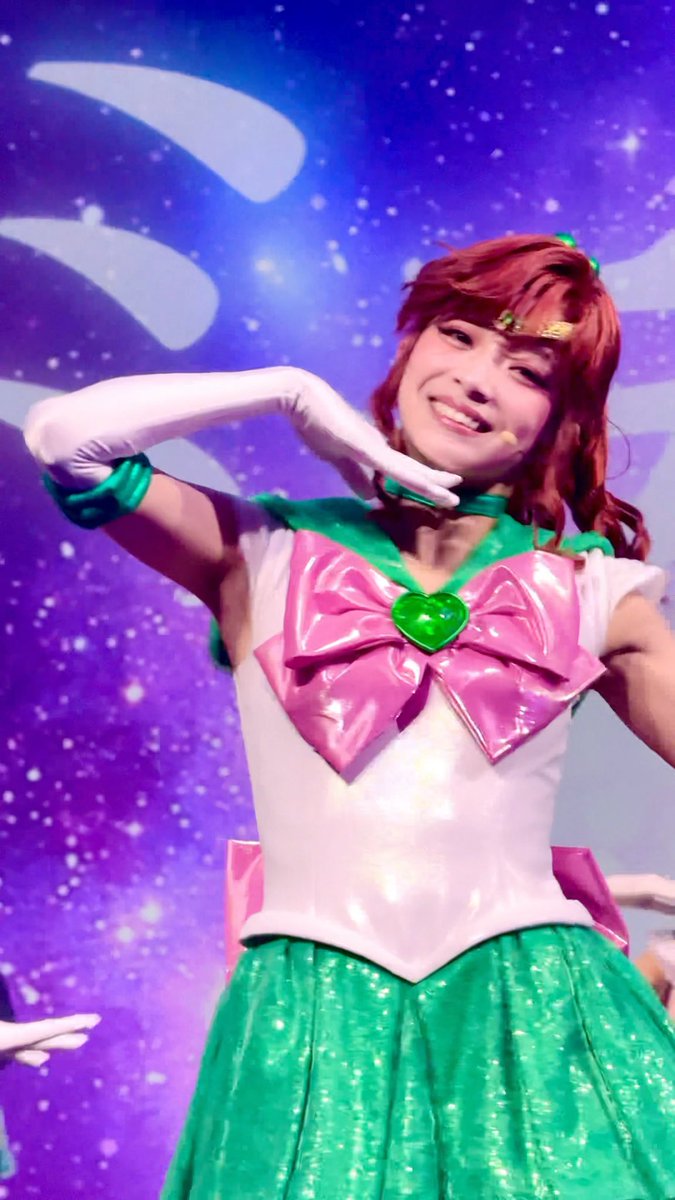 A performer with red hair, wearing a pink bow and green skirt Sailor Jupiter-style costume, smiles and strikes a pose on stage against a starry purple backdrop.
