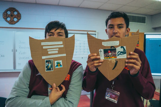 Two individuals in a classroom hold up cardboard shields with cut-out photos and text. The shields display an image of Jesus and a photo of a dolphin among other images. A whiteboard and classroom items are visible in the background.