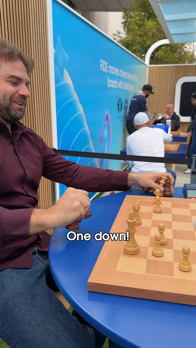 A man in a maroon shirt sits at a blue table playing chess, holding captured pieces in his fist while the caption "One down!" appears on screen. In the background, a banner reads "FIDE moves chess beyond the board with Salesforce," capturing a candid moment from FIDE's chess activation at Dreamforce where players competed in matches and puzzles.