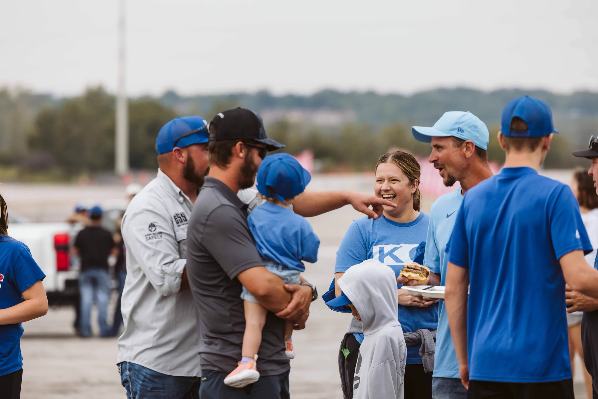 A group of people dressed in blue and casual attire are socializing outdoors. Some are wearing baseball caps. One person is holding a small child. They're engaged in conversation with visible smiles. A greenery-filled background and overcast sky are visible.