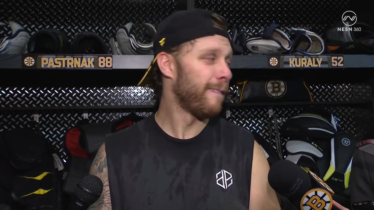 David Pastrňák (#88), wearing a black cap backward and a black tank top, smiles in the Boston Bruins locker room during an interview, with nameplates for "PASTRNAK 88" and "KURALY 52" and Bruins gear visible behind him — fitting the post about the Bruins' season win song "Unwritten" by Natasha Bedingfield, which Pasta is a big fan of.