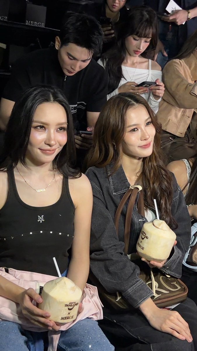 Two young women smile at the camera while sitting in a crowd; they are holding coconuts branded with 'THAILAND x MERCE' and are accompanied by people in the background.