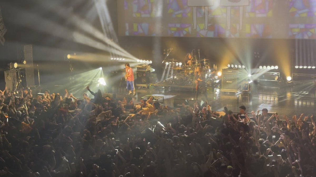 A packed concert venue is illuminated by dramatic stage lights as a band performs, with the crowd enthusiastically raising their hands high in the air—capturing the exhilarating, communal energy described in the caption about the awe-inspiring sea of raised hands.