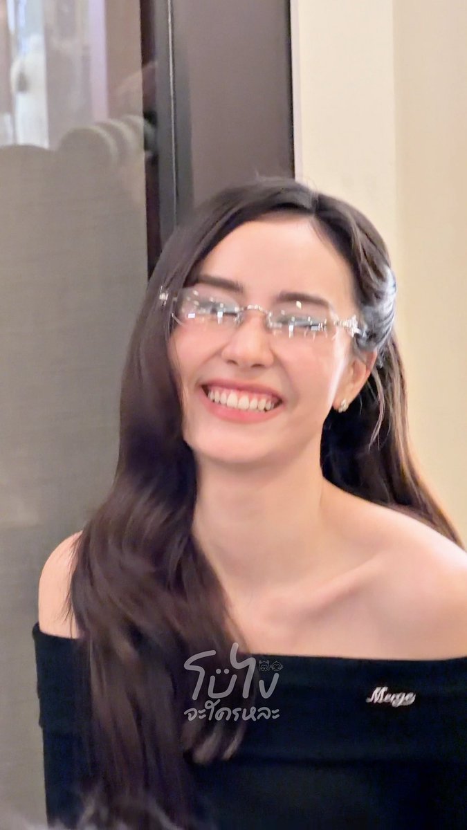 A smiling woman with long brown hair wears rimless glasses and an off-the-shoulder black top. Thai text 'โฆษง จะใครนะ' appears at the bottom.