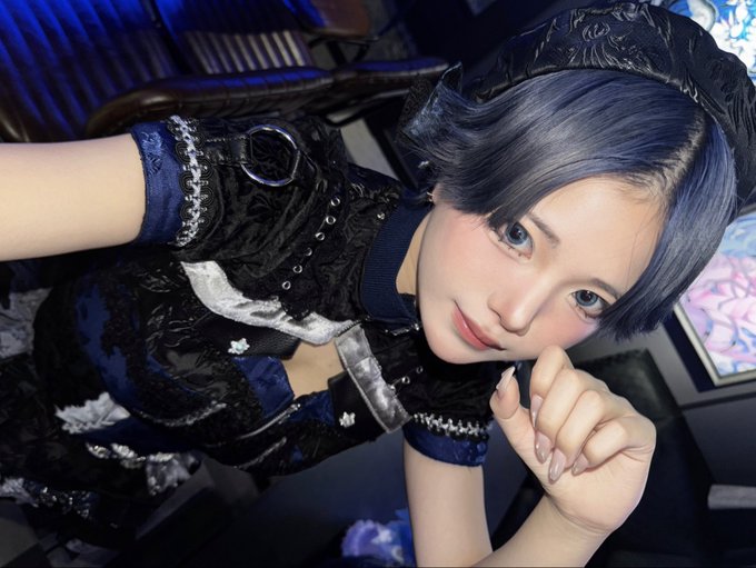 A close-up selfie of a young woman with short dark hair wearing an ornate dark blue and black dress with lace and silver details, resting her chin on her hand.