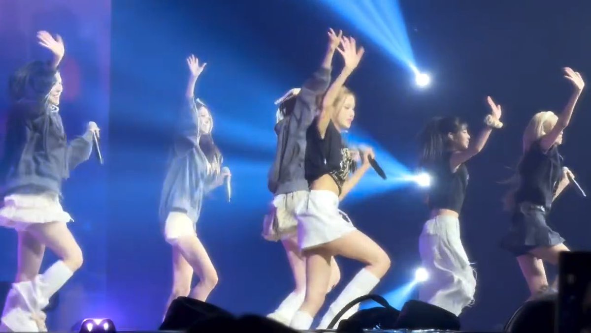 K-pop girl group performing on stage with blue lighting, members wearing matching outfits with skirts, white knee-high boots, and holding microphones while dancing with raised arms.
