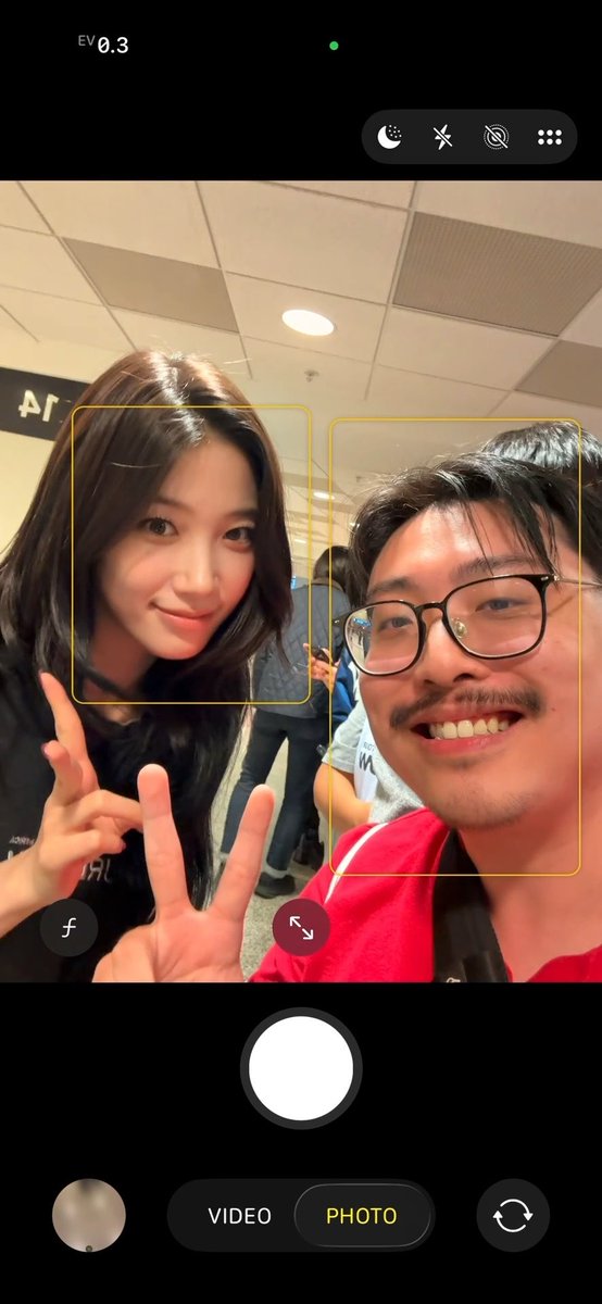 A screenshot of an iPhone camera interface showing a young woman and a man with glasses posing for a photo, both detected as faces by the camera's AI.