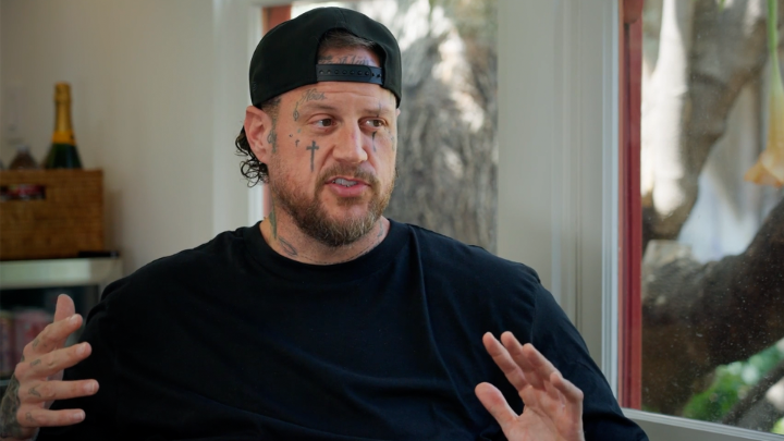A man wearing a black backward baseball cap and black shirt speaks with his hands raised expressively; he has visible face tattoos including a cross under his eye.